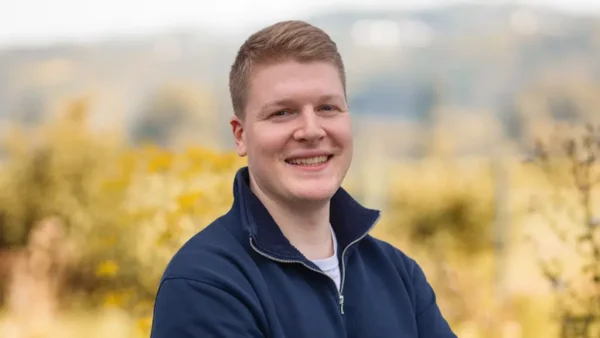 Simon Geib, freundlicher Ansprechpartner bei eventflotte.de, lächelnd vor unscharfem Naturhintergrund mit gelben Farbtönen.